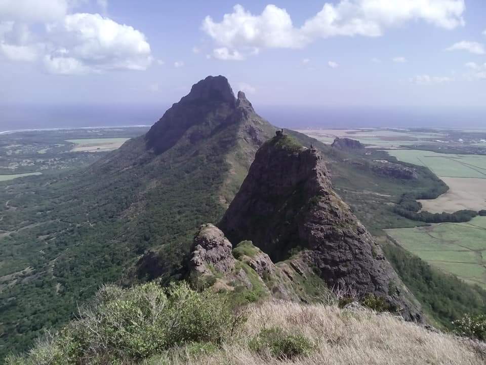 View from mountain top of Mauritius showing triangular top of rocky grey mountain and 2 further peaks underneath a blue sky.