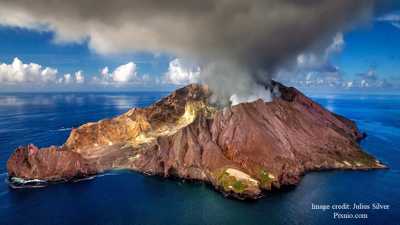 Small grey and rugged volcano in the ocean spewing out grey clouds of volcanic materials.