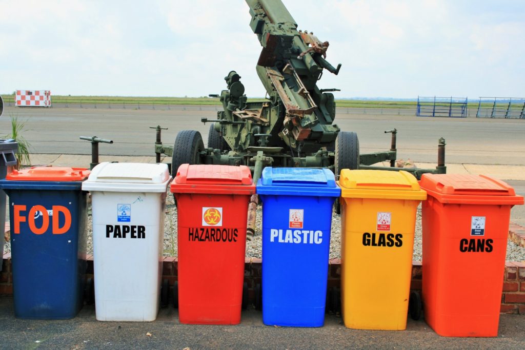 6 different bins for recycling, from left to right, dark blue bin with FOD written on it, grey bin paper written on it, red bin hazardous written on it, light blue bin plastic written on it, yellow bin glass written on it and orange bin cans written on it