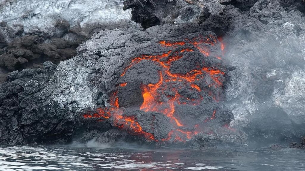 Orange lava enveloped in grey rock falling into ocean thus causing tsunamis, high waves