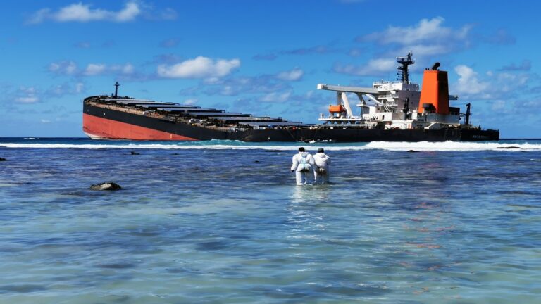 MV Wakashio oil spill in Mauritius; two people in white overall standing in the sea with black and red ship tilted in the back on coral reef
