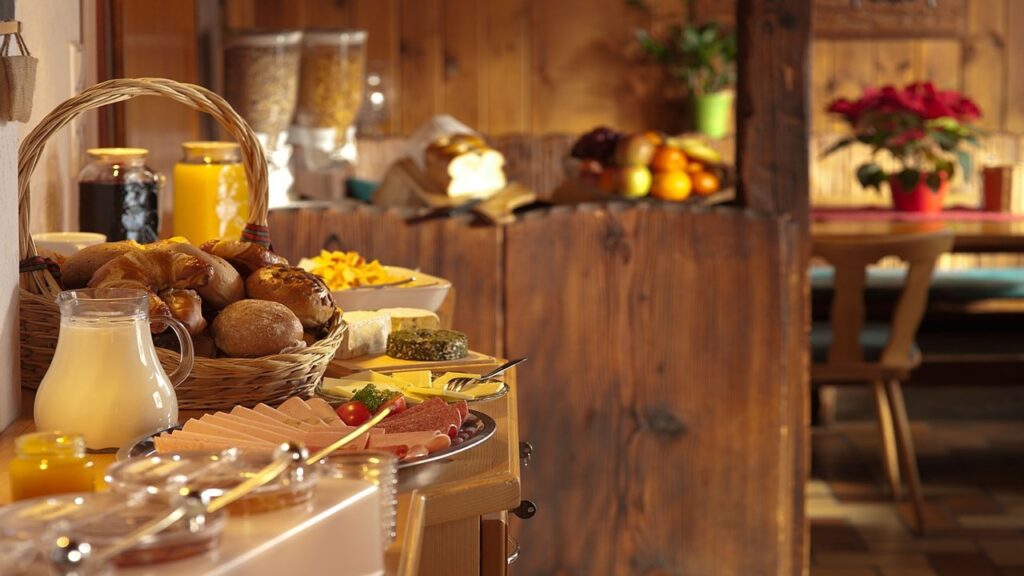 Difference between sustainable tourism, ecotourism and green tourism; wooden kitchen with food on brown wooden table, white milk jug, yellow orange juice, cream sausage and fruits
