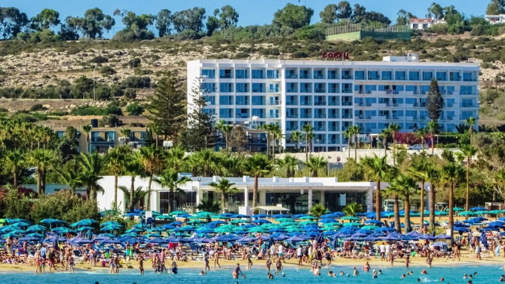 Mas tourism at the beach, many people and blue umbrellas on white beach in front of white hotel and green trees; negative effects of tourism on the environment