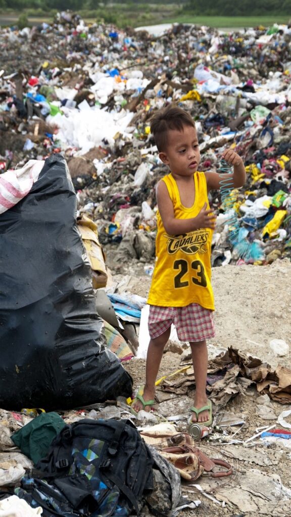 Small child wearing yellow shirt and striped red shorts playing with trash in trash filled place, green living