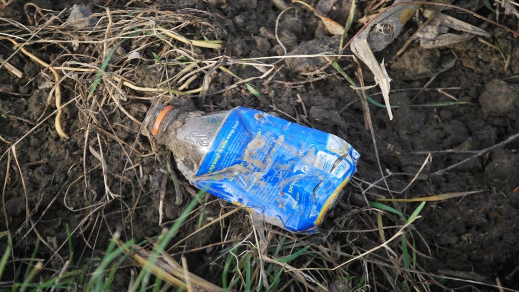 Clear bottle with blue cover lying on brown ground and green grass, plastic pollution