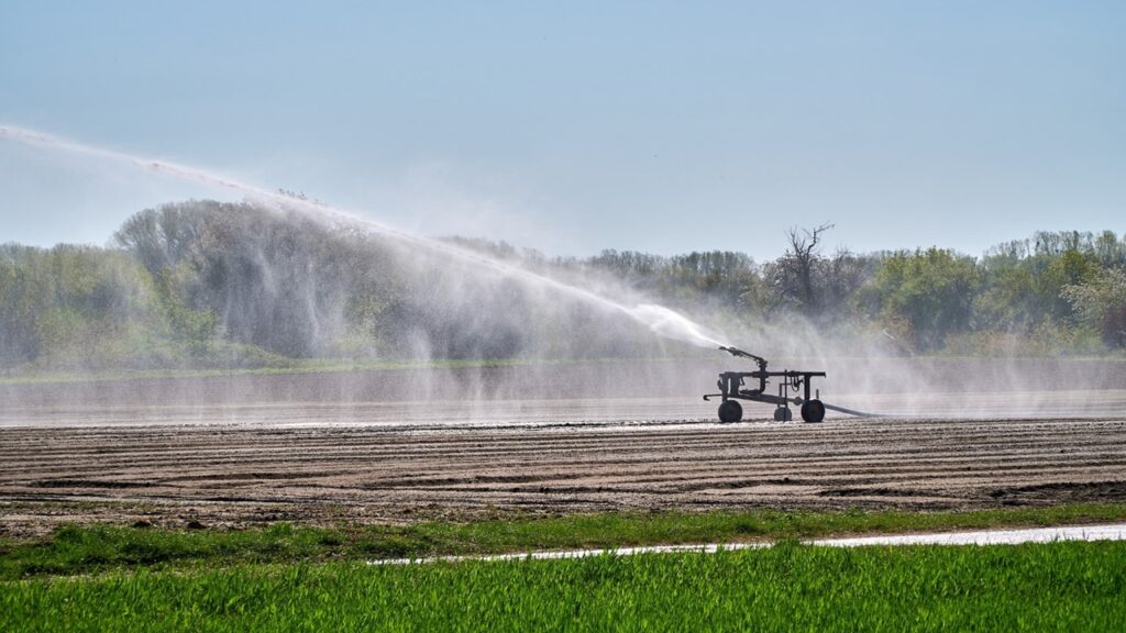 Machine irrigating brown land in green field over blue sky with white water splashes